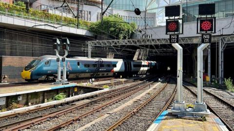 A blue and yellow train is pulling into a platform on the left. There are tracks and signals in foreground and bridge and buildings in the distance.