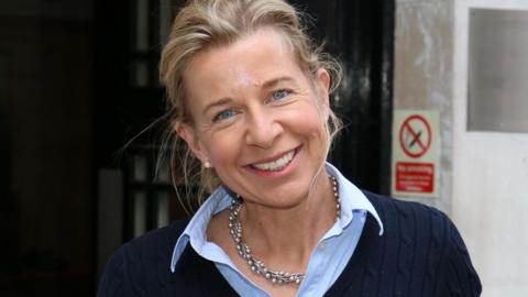Katie Hopkins smiles at the camera with her head tilted to her right shoulder. She is wearing a navy blue jumper with a blue shirt underneath.