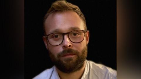 The head and shoulders of Matt Burgin-Powell against a dark background. He is wearing an Oxford-style blue cotton shirt, has a thick brown beard, short hair with a quiff, and glasses.