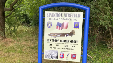 The sign for Spanhoe Airfield. The sing is held in a bright blue metal frame. The sign says "Spanhoe Airfield USAAF station 493". It has a picture of what looks like a Dakota aircraft, used during World War Two.