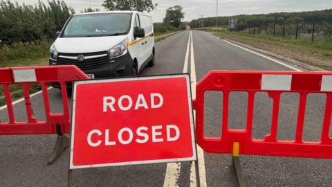 A red 'ROAD CLOSED' sign and red barriers blocking a road; a white van is parked behind the barriers.