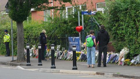 Four people stand in front of a row of floral tributes outside the school. There are lots of bunches of flowers and one red heart balloon. 