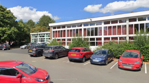 Image taken in the car park of the library with several cars parked up in spaces in front of the main building. The building is white with large rectangle windows and red curtains hanging inside the windows. 