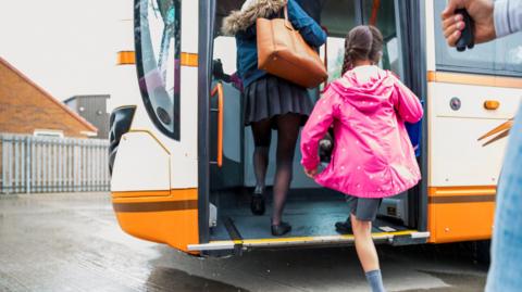 Two girls with their backs to the camera are getting on their school bus. The girls are both wearing grey skirts and coats. The bus is painted in a mixture of orange and white