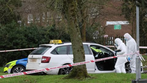 A white estate car with a taxi sign sits in a car park. There is a police cordon tied to a street sign. A marked police car is parked behind the taxi. There are a variety of trees and patches of grass at the perimeter. Two forensic officers in white body suits examine the taxi.