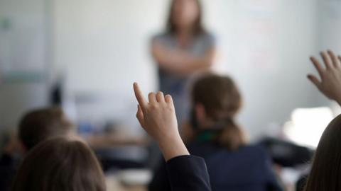 A raised hand, with a raised index finger, is the only clear object in an otherwise blurry picture. However, the scene shows a classroom environment in which we can see the back of seated pupils' heads and a teachers standing, arms folded, at the front of the class. 