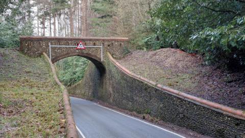 A stone bridge in a countryside. The bridge stands over a tarmac road with high walls leading up to it.