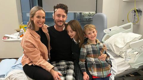 Matthew White wearing pyjamas and sitting on a hospital bed, with his wife on his right hand side and his son and daughter on his left.