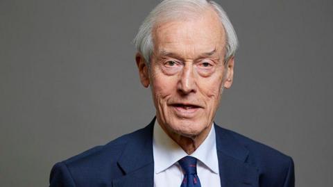 A head and shoulders picture of a man wearing a blue suit, a blue tie and a white shirt. He has grey hair and is smiling at the camera.