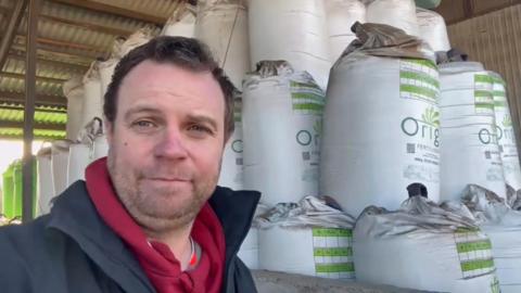 Olly Harrison has short brown hair, he is wearing a red hooded top and a blue jacket and is looking at the camera while stood in front of large stock of fertiliser