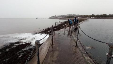 The wall between Clevedon Marine Lake and the sea next to it. People are on the wall cleaning it. They are wearing blue high vis vests.