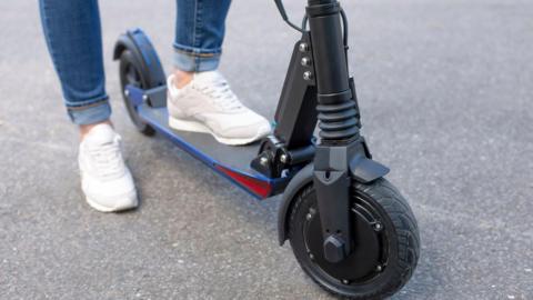 A close-up of a person wearing blue jeans and white trainers with their foot on an e-scooter on the pavement.