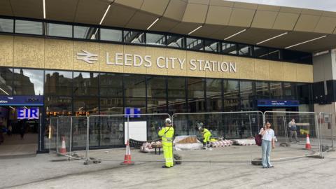 The front of Leeds railway station. There is a gold sign above a glass frontage, with the words "Leeds City Station" in white. There are safety barriers in front of the windows, and workers in hi-vis clothing can be seen. Some passengers are also standing around the entrance.