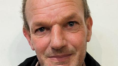 A close-up of a Nick Starmer, who has light skin and short hair that is thinning at the top. His expression is neutral but slightly warm, with a faint smile. The background is plain and white. He is wearing a dark jacket and a light-coloured shirt.