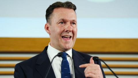 Russell Findlay, who has short black hair, speaks into a microphone. He is wearing a business suit