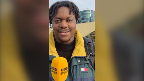 A man with black hair wears a green jacket with a yellow collar and a BBC Sport microphone.