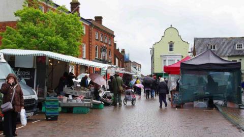 People visiting stalls set up at Fakenham's Market Place on a rainy day. Striped canvas coverings and pop-up marquees are covering the stalls.