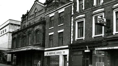 A black and white photograph of the front of the theatre building. A white sign with black writing says Court Cinema.