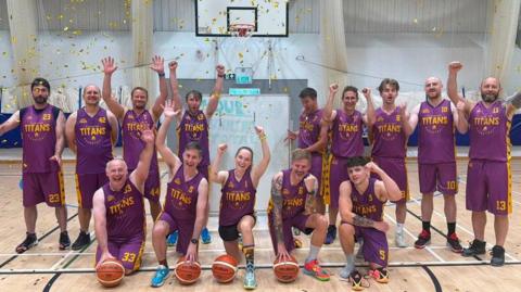 A line up of Tiverton Titans basketball players on a court. They are all smiling and wearing purple short-sleeved tops and shorts with the word Titans in yellow.