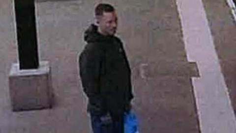 Grainy CCTV still of a man with short brown hair, a green coat carrying a blue plastic bag
