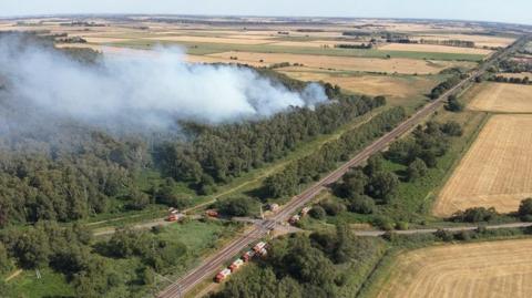 An aerial image of smoke rising out of woodland. A railway line is next to the trees, with a minor road crossing the line.