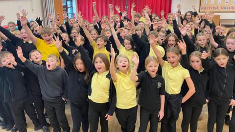 A large group of schoolchildren in black uniforms with yellow polo shirts all have one hand in the air and are looking towards the camera. Their faces show they are singing. They are indoors in a hall with red curtains in the background and a noticeboard that says British Values on it.
