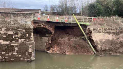 Traffic cones line a section of road across Old Powick Bridge where the supporting wall is completely missing. Bare earth can be seen where the wall has been swept away