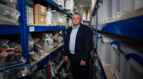 Det Ch Insp Paul Curtis stands facing the camera in the aisle of the evidence room, piled high with luxury goods seized from financial fraudsters
