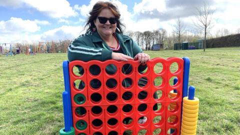 A woman has shoulder length auburn hair and is wearing a green tracksuit top with white stripes down the arm. She has sunglasses on and is smiling as she leans over a giant Connect 4 game