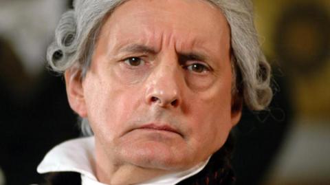 A man in a powdered wig and 18th-Century costume looks soulfully into the camera. 