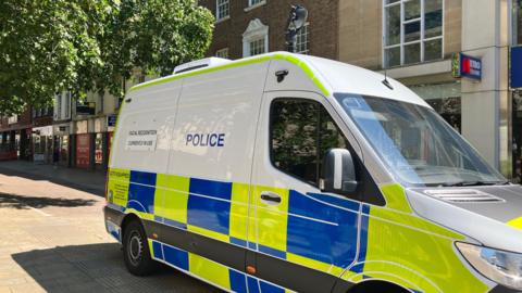 A white police van with green and blue checks on it and black cameras on the roof.