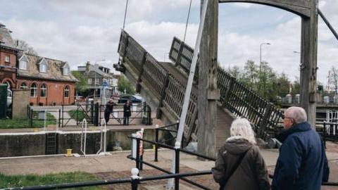 A wooden footbridge crossing Mutford Lock being lifted with pedestrians watching on either side   