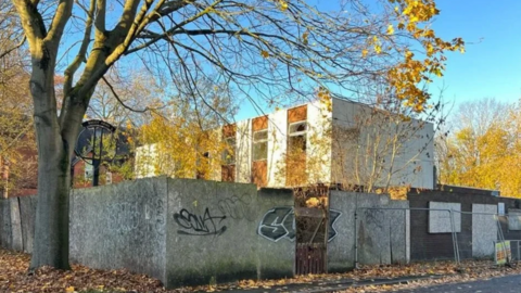 The pub is surrounded by boards to close it off, some have graffiti on them. The pub can be seen behind the boards and is white and brown with windows. A large tree stand in front and has shed its autumn leaves on the ground.