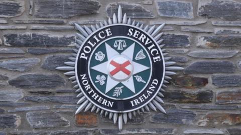 A green, black, grey and red PSNI badge sits on a stone wall. The writing on the crest reads ' Police Service of Northern Ireland'.
