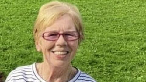 Una Noone is smiling into the camera. She has glasses on and a black and white stripped top. She had short blond hair. She's standing in a field. 