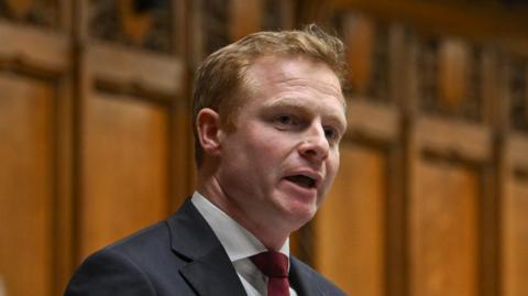 A man with short ginger hair wearing a navy suit and burgundy tie speaks in the House of Commons.