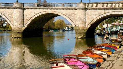 Sunshine reflecting off the water and bridge with a line of colourful row boats lined up on the bank