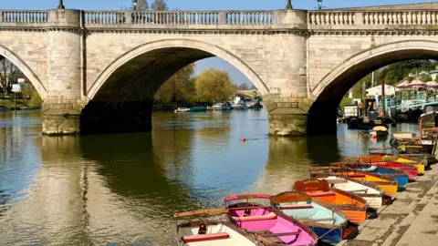 Sunshine reflecting off the water and bridge with a line of colourful row boats lined up on the bank