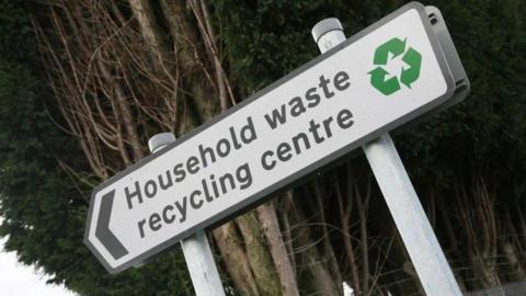 An arrowed sign with the words "Household waste recycling centre", alongside a green recycling logo.