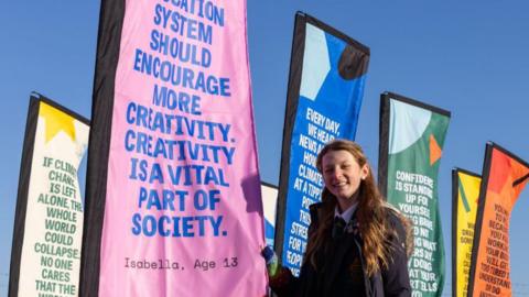 A young girl in a school uniform standing in front of colourful flags displaying inspirational messages. The pink flag closest to her reads, "The education system should encourage more creativity. Creativity is a vital part of society." The sky is clear and blue.