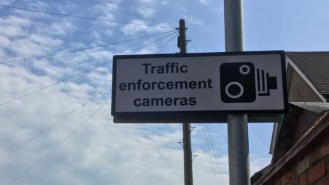 White sign on a pole, with black lettering "Traffic Enforcement Cameras" and a camera symbol. A house roof and the sky are visible behind.