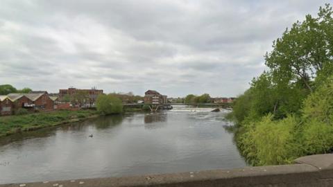 A canal, pictured from a bridge above. Trees and industrial buildings can be seen flanking the water.