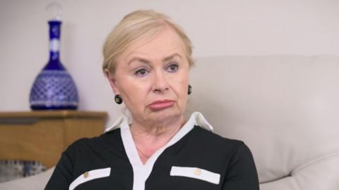 A senior woman sits on a sofa being interviewed.