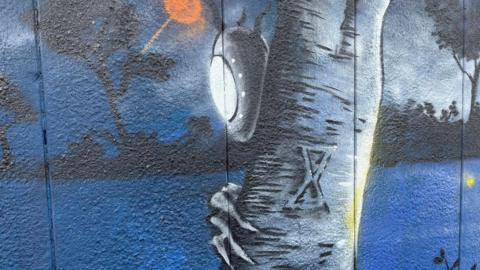 A spray paint artwork on a wall, showing an alien with a lit-up mask and grey hand peering round the trunk of a tree. It is depicted at night time, with lots of dark blue in the background and the silhouettes of trees.