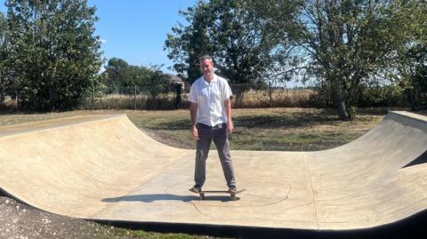 Mark Ormston is trying out the new skate park. He is standing in the dip of the ramp on top of a skateboard with his arms down by his sides. He is smiling while wearing grey jeans and a white shirt. It is a sunny day with blue skies.
