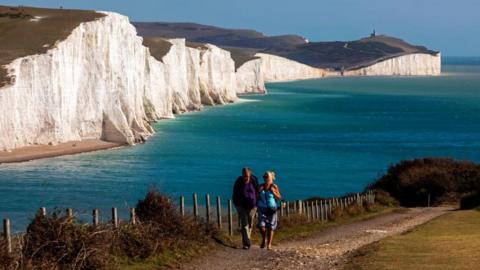 The white Seven Sisters cliffs are visible in the background, with the blue water of the Channel. A man and a woman dressed in hiking clothes are walking up a path on the right of the shot