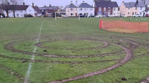 Circles of muddy tyre tracks left by a car, most likely doing doughnuts on the field. There is a football goal in the background and a row of houses behind it. Part of the pitch has already been fenced off by orange netting.