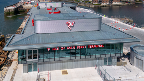 Aerial shot of the front of the ferry terminal, which has Isle of Man Ferry Terminal written across the windows on the second floor and a Manx triskelion on the roof above. There is water either side and red brick building to the right behind it.