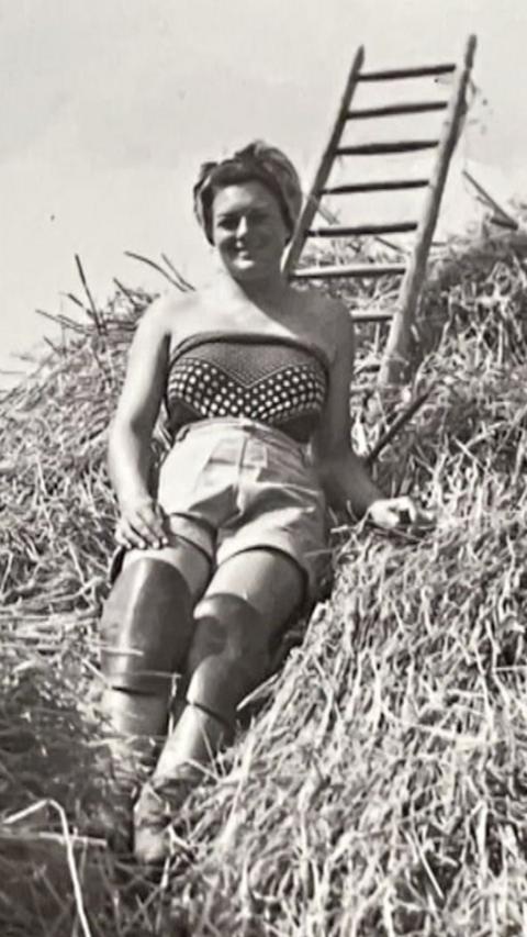 A black and white image of a young Gwen Raggett leaning against a thatched roof. She wears a silk scarf as a strapless top with shorts and leather knee pads on and her hair is wrapped in a headscarf.