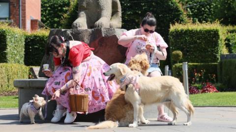 Two women dressed in pink and both with dark hair and sun glasses are with three light-coloured dogs. They are feeding the dogs ice cream